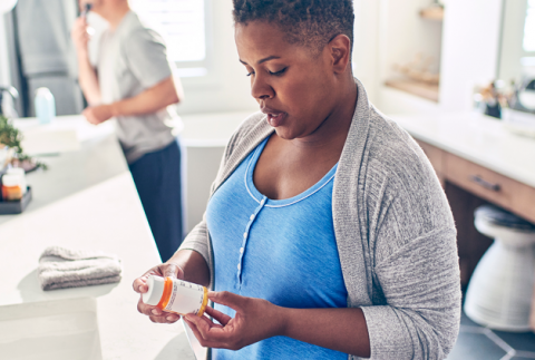 Black female in bathroom reading the label of a medication bottle from ESC's online pharmacy while man is shaving in the background.